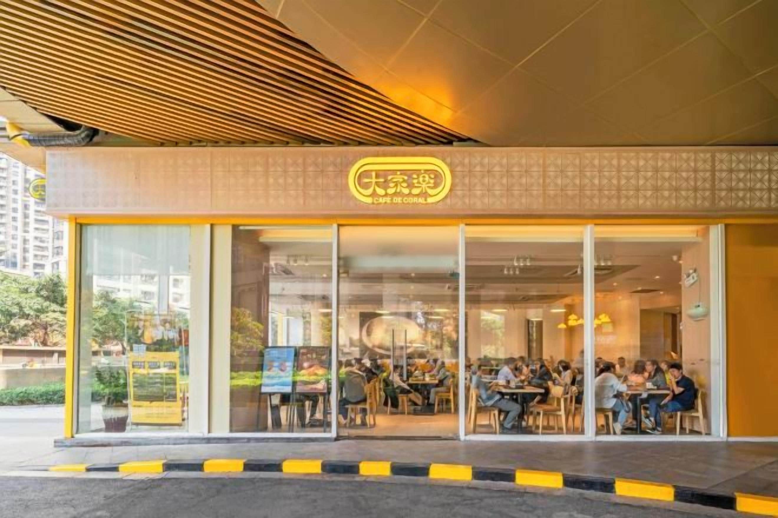 Una cafetería con fachada de vidrio, mesas al aire libre y un letrero amarillo.