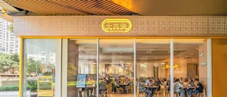 Una cafetería con fachada de vidrio, mesas al aire libre y un letrero amarillo.