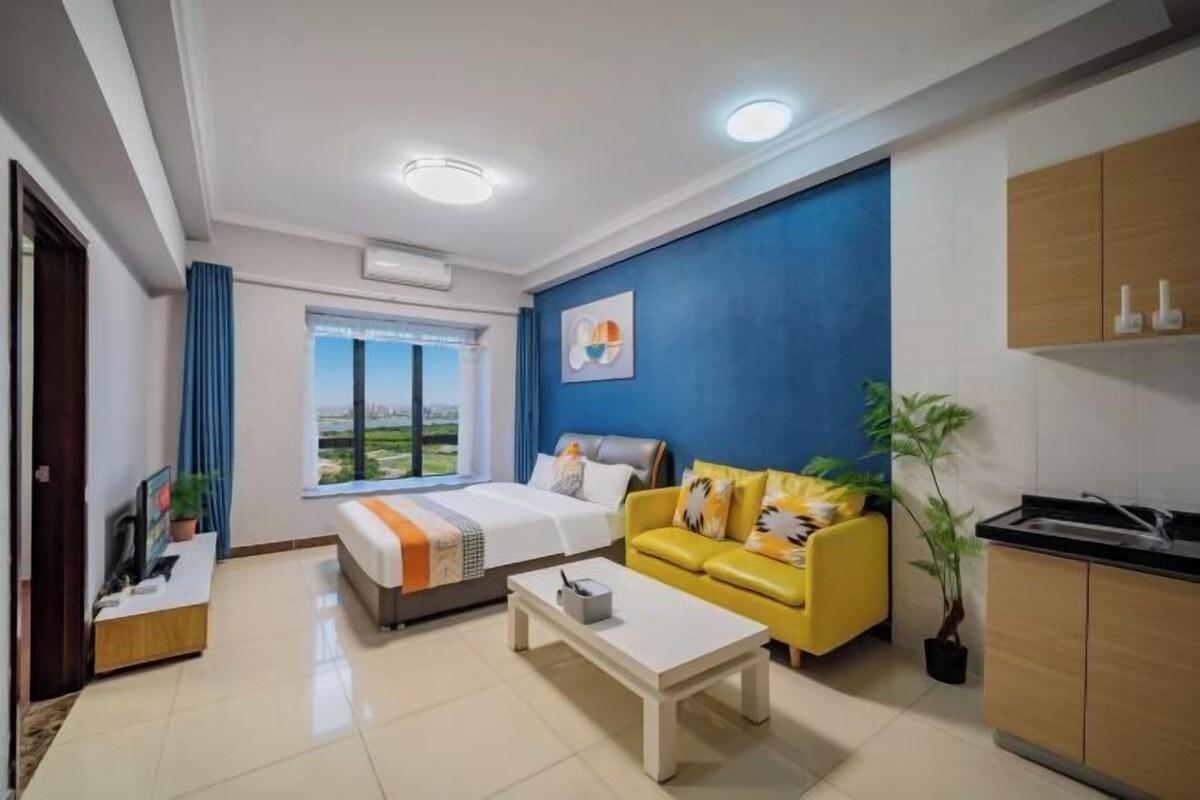 A modern hotel room with a bed, a yellow sofa, a coffee table, a kitchen area, and a large window.