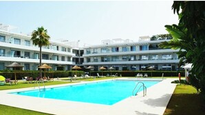 Piscina cercada por espreguiçadeiras e guarda-sóis, com um edifício alto ao fundo.