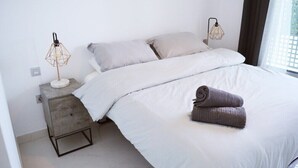 Uma cama bem arrumada com roupa de cama branca e duas almofadas, uma pequena mesa de cabeceira com abajur e uma janela com cortinas.