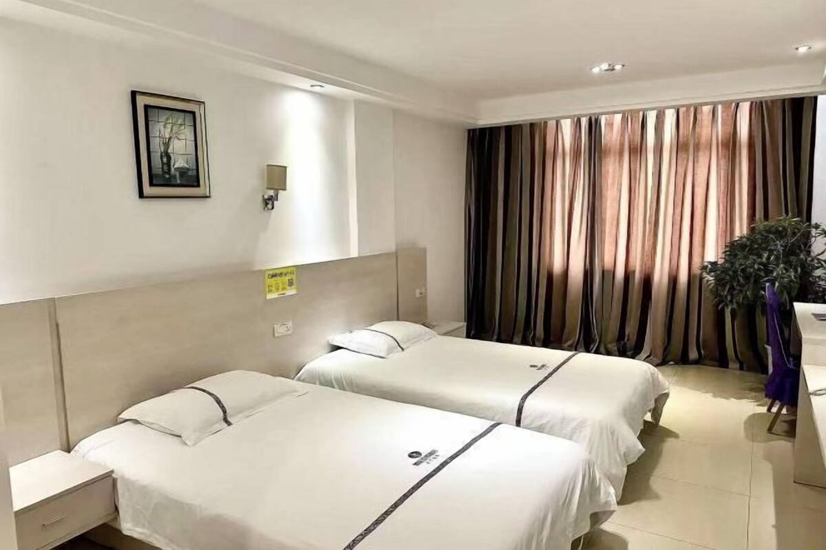 A hotel room with two beds, a nightstand, a wall-mounted light, a framed picture, and a curtain.