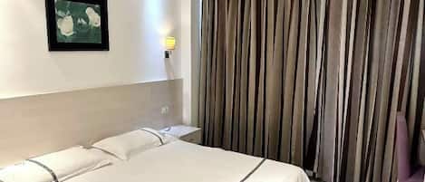 A hotel room with a bed, a nightstand, a wall-mounted lamp, a framed picture, and a curtain.