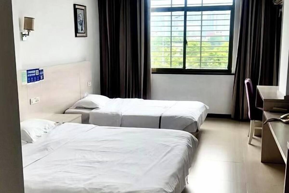 A hotel room with two beds, a desk, a chair, a window with curtains, and a wall-mounted lamp.