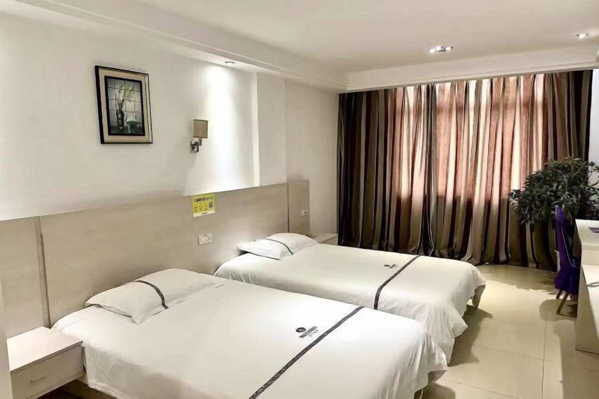 A hotel room with two beds, a nightstand, a wall-mounted light, a framed picture, and a curtain.