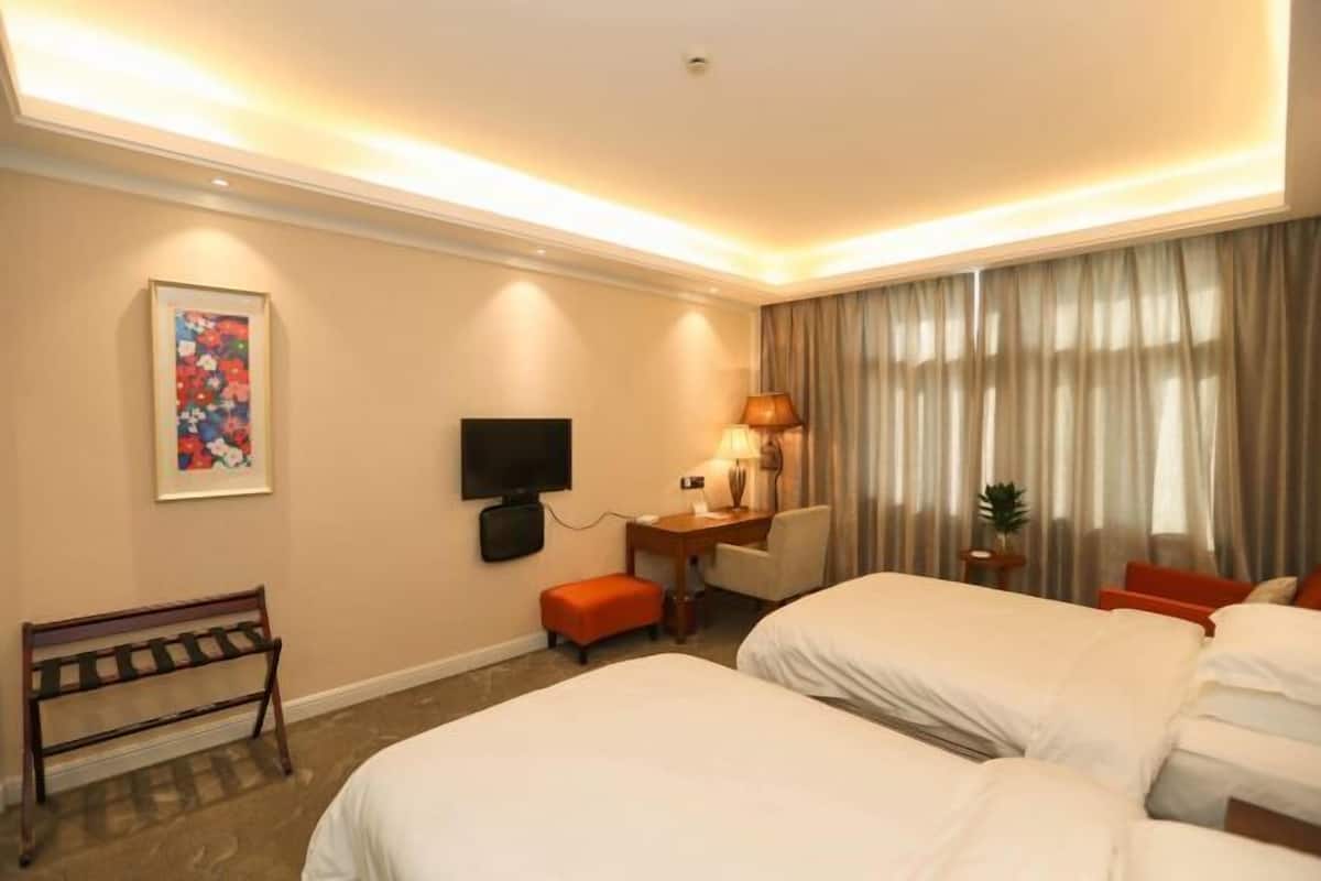 Une chambre d’hôtel avec deux lits, un bureau avec une chaise, une télévision, un banc et une fenêtre avec des rideaux.