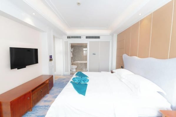Une chambre d’hôtel avec un grand lit, un téléviseur fixé au mur, un banc en bois et une salle de bain visible à travers une porte ouverte.