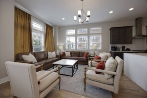 A spacious living room with a brown leather sofa, beige armchairs, and a coffee table. There are large windows with curtains, a chandelier, and a kitchen area visible.