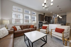 Een woonkamer met een bruine leren bank, een witte salontafel en twee witte fauteuils met rode kussens. Achtergrond toont een keuken met donkere kastjes en een wit aanrecht.