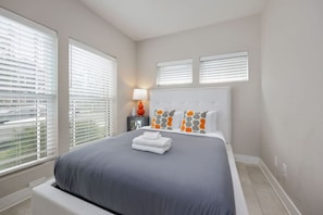 Un dormitorio con una cama grande, cabecera blanca y dos ventanas con persianas.