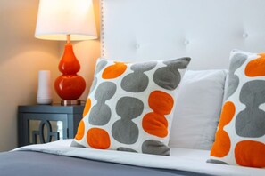 A bed with two pillows featuring a polka dot pattern in orange and gray, a white headboard, and a nightstand with a red lamp and a white object.