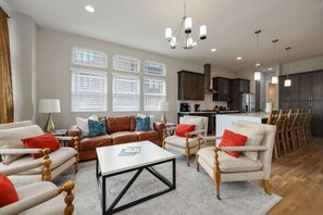 Un salón espacioso con un sofá de cuero marrón, sillones blancos y una mesa de centro. Se observan varias ventanas con persianas, un candelabro y una zona de cocina al fondo.