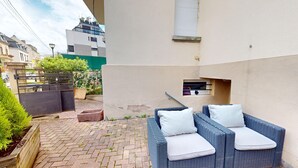 Een klein terras buiten met rieten meubels en kussens, een pad van bakstenen en een groene omheining.