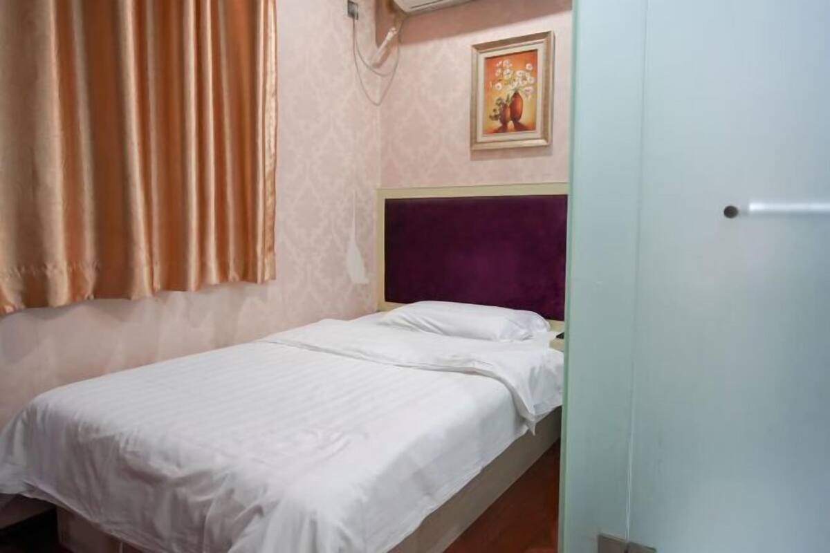 Una cama individual con colcha blanca, cabecera morada y aire acondicionado de pared.