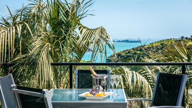 Una mesa de vidrio con una botella, un cuenco y un plato, rodeada de sillas y con vistas a un paisaje con palmeras y una masa de agua.