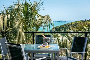 Une table en verre, sur laquelle se trouvent une bouteille, un bol et une assiette, est entourée de chaises et offre une vue panoramique sur un paysage agrémenté de palmiers et d’une étendue d’eau.