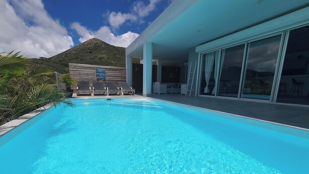 A clear blue pool with lounge chairs, a mountain in the background, and a modern building with large windows.