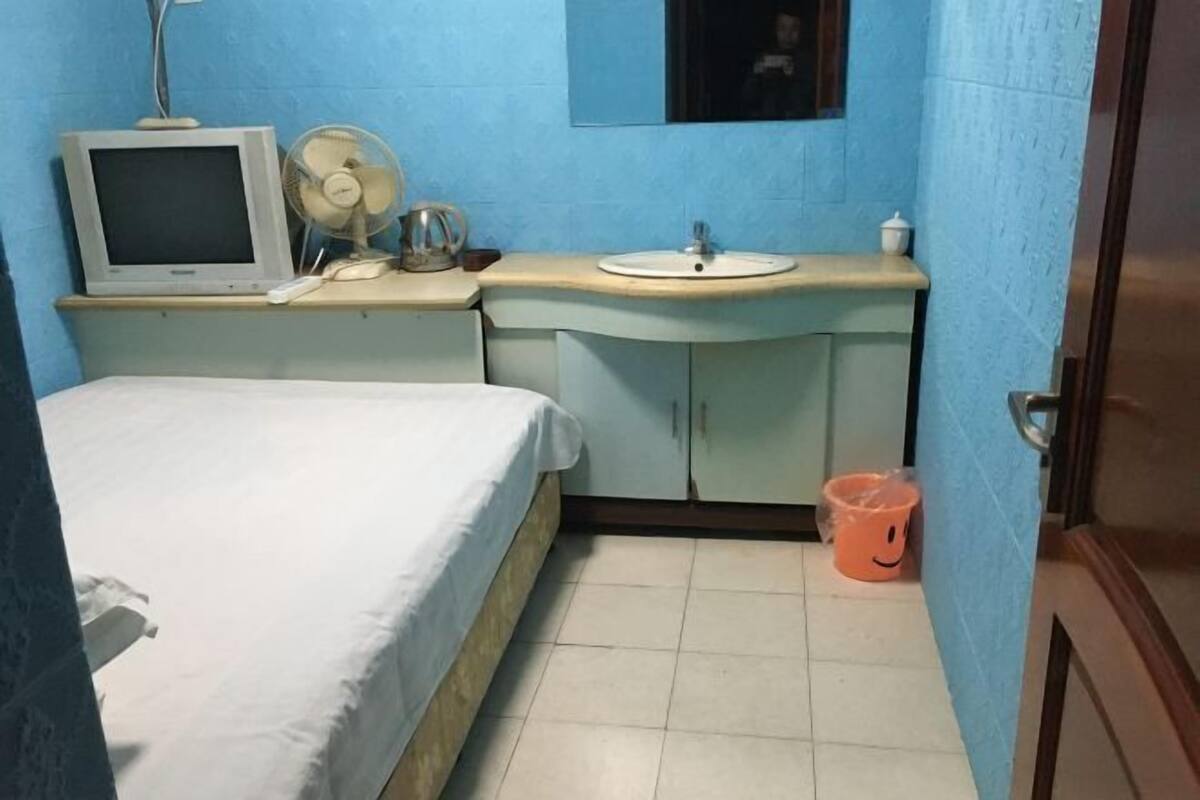 Ein Zimmer mit einem Bett, einem kleinen Schreibtisch mit Computerbildschirm, einem Ventilator, einem Waschbecken und einem Mülleimer.