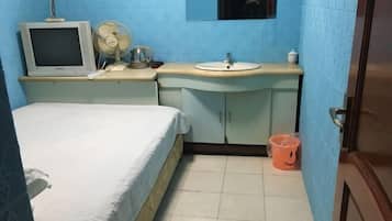 Una habitación con una cama, un escritorio pequeño con un monitor de computadora, un ventilador, un lavamanos y un cubo de basura.