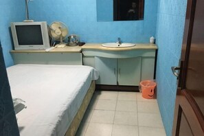 Una habitación con una cama, un escritorio pequeño con un monitor de computadora, un ventilador, un lavamanos y un cubo de basura.