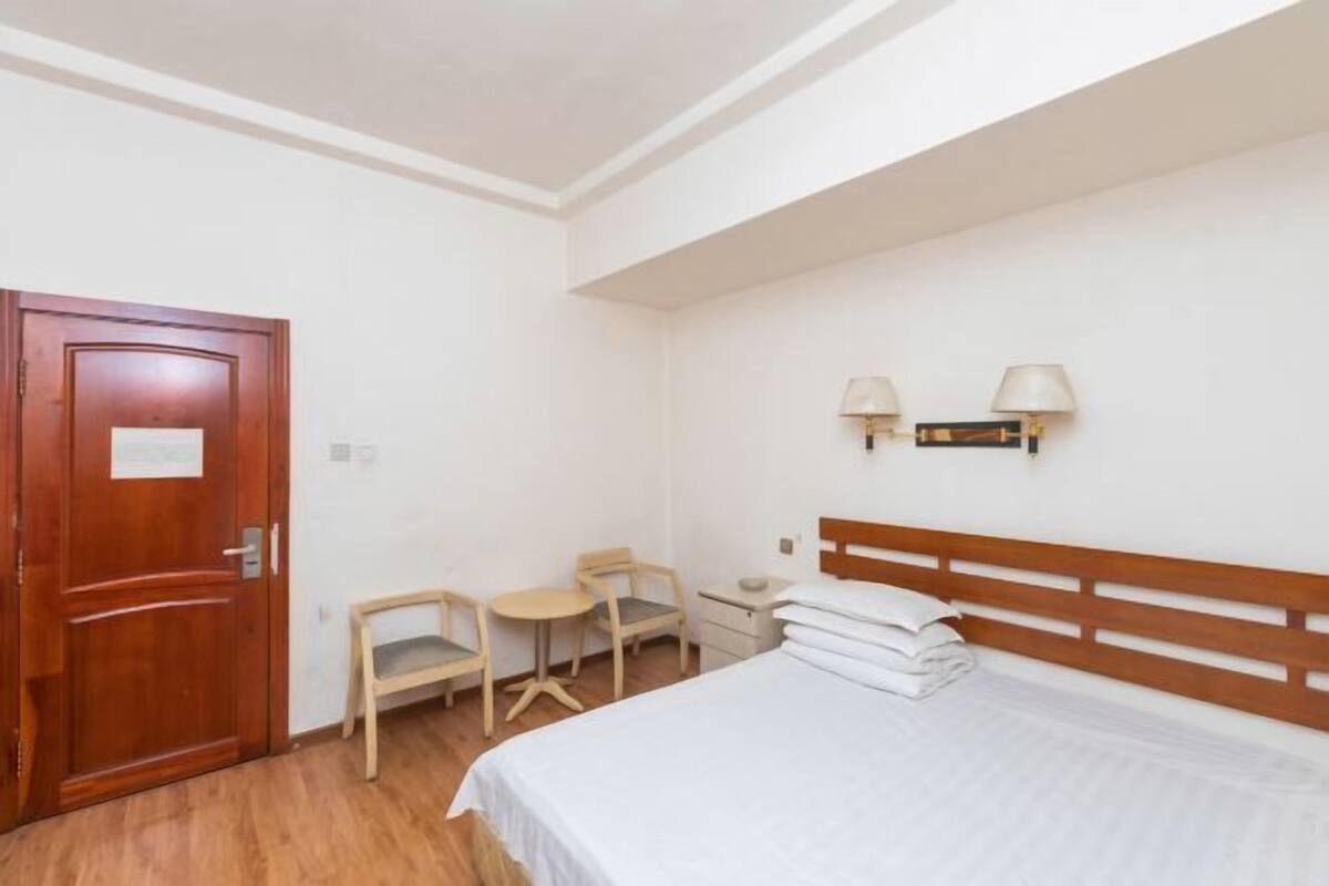 Ein Schlafzimmer mit Holztür, einem Bett mit weißer Bettwäsche, zwei Nachttischlampen, einem kleinen Tisch mit Stuhl und einem hölzernen Kopfteil.