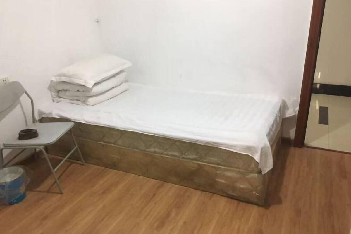 Ein kleines Zimmer mit einem Einzelbett, Holzboden, einem Metallstuhl und einem kleinen Tisch.