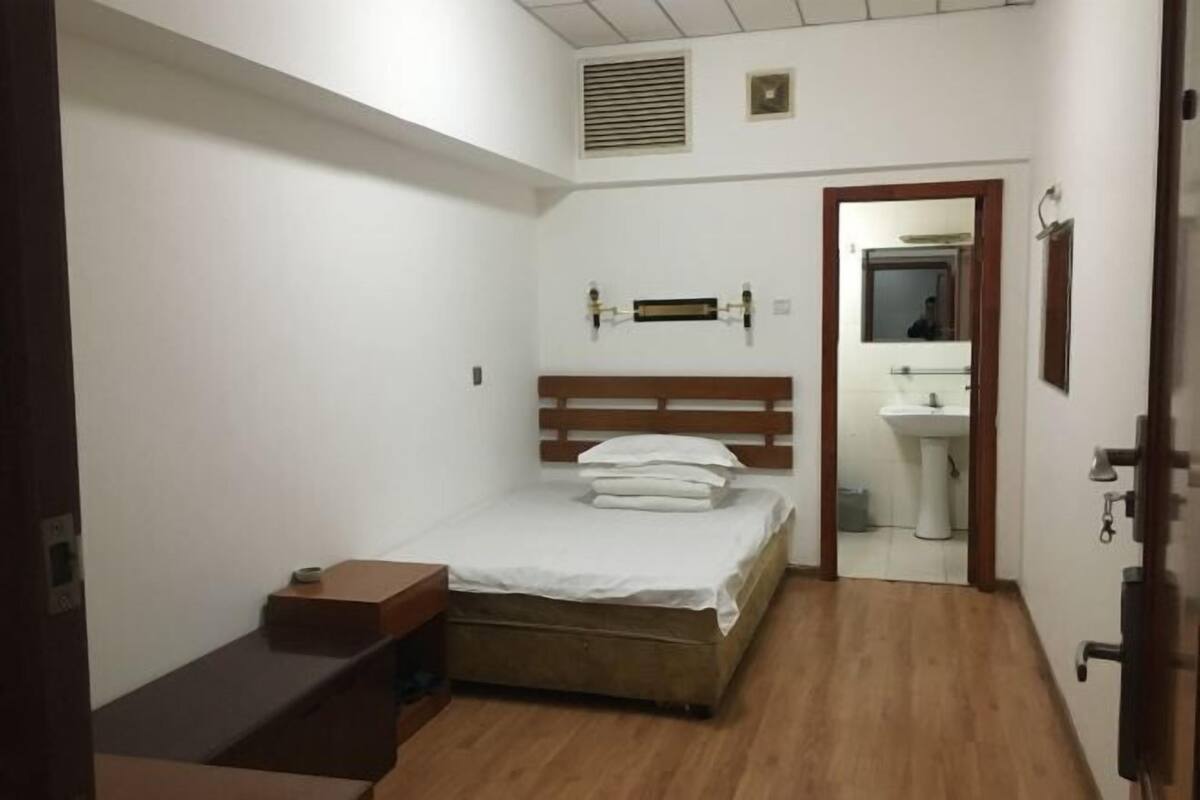 Ein kleiner, sauberer Raum mit einem Einzelbett, einer hölzernen Sitzbank, einem Nachttisch, einem Spiegel und einem Waschbecken im anliegenden Badezimmer.