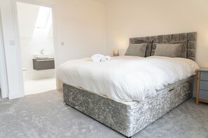 A bedroom with a bed, a nightstand, a lamp, and a bathroom in the background. - 3 Bedroom, 3.5 Bathroom House, Goodwood, Chichester (Chichester)