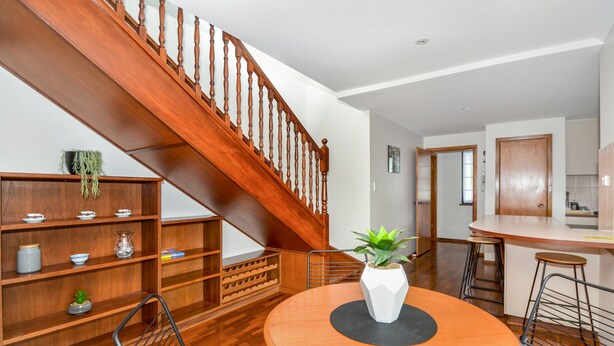 Un coin repas avec un escalier en bois, une table ronde entourée de chaises et, au fond, une cuisine.