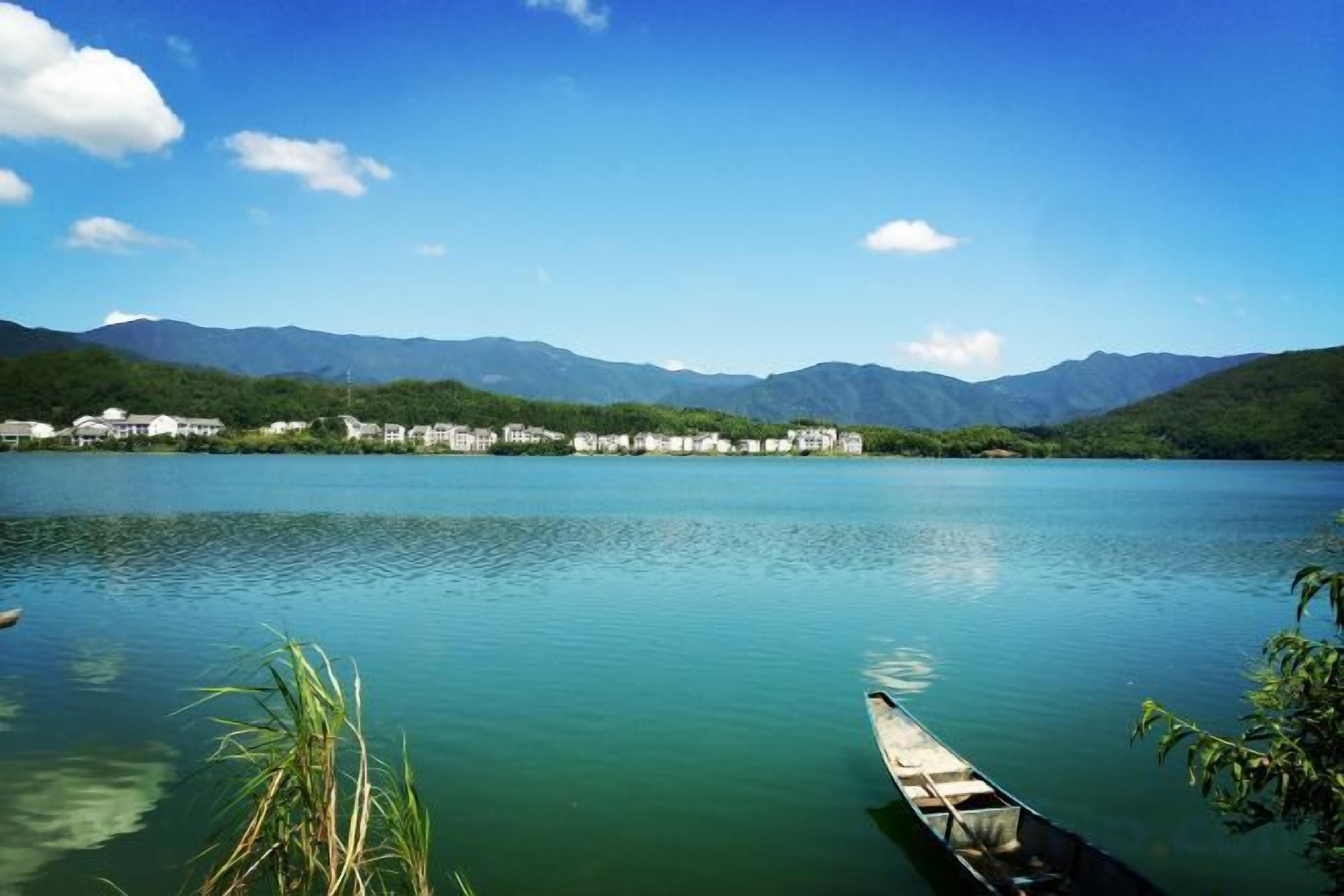 A calm lake with a wooden boat docked on the shore, surrounded by mountains and buildings in the background.
