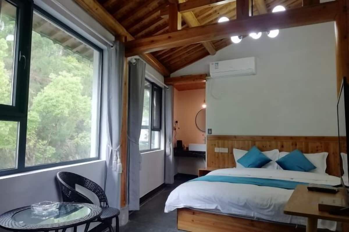 Habitación con cama, una mesa y una silla. Se ve un ventanal con vista a árboles.