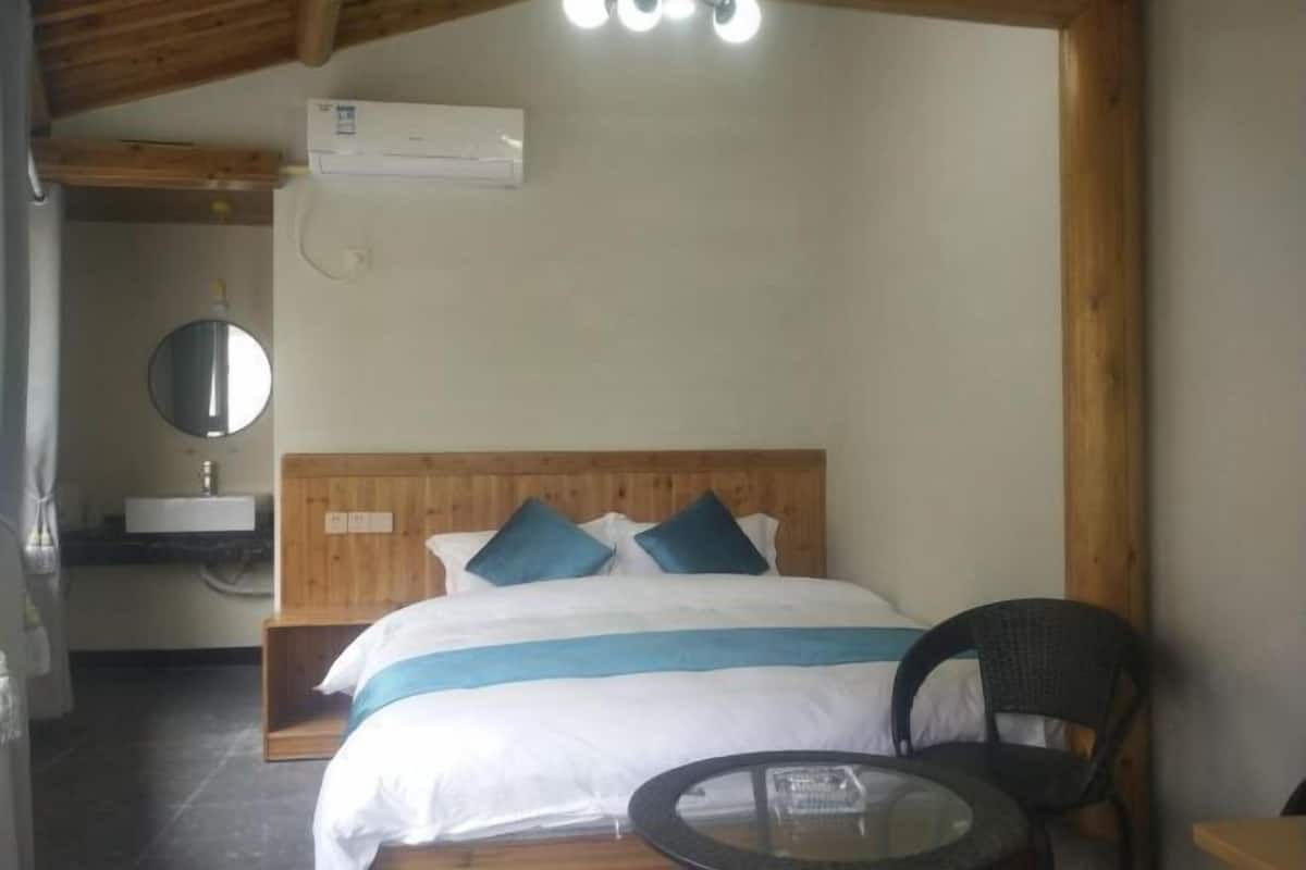 Un dormitorio con cabecera de madera, cama con ropa blanca y detalles azules, un espejo redondo, una mesita pequeña y una silla.