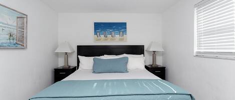 Un dormitorio ordenado con cama, dos mesitas de noche con lámparas, un ventilador de techo, un cuadro con motivos playeros y una ventana con persianas.