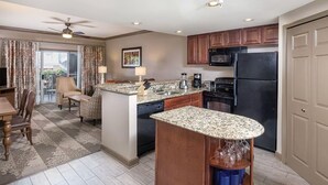 Una cocina moderna con encimeras de granito, armarios de madera y un refrigerador negro. Hay un comedor con una mesa y sillas, y un balcón con cortinas.