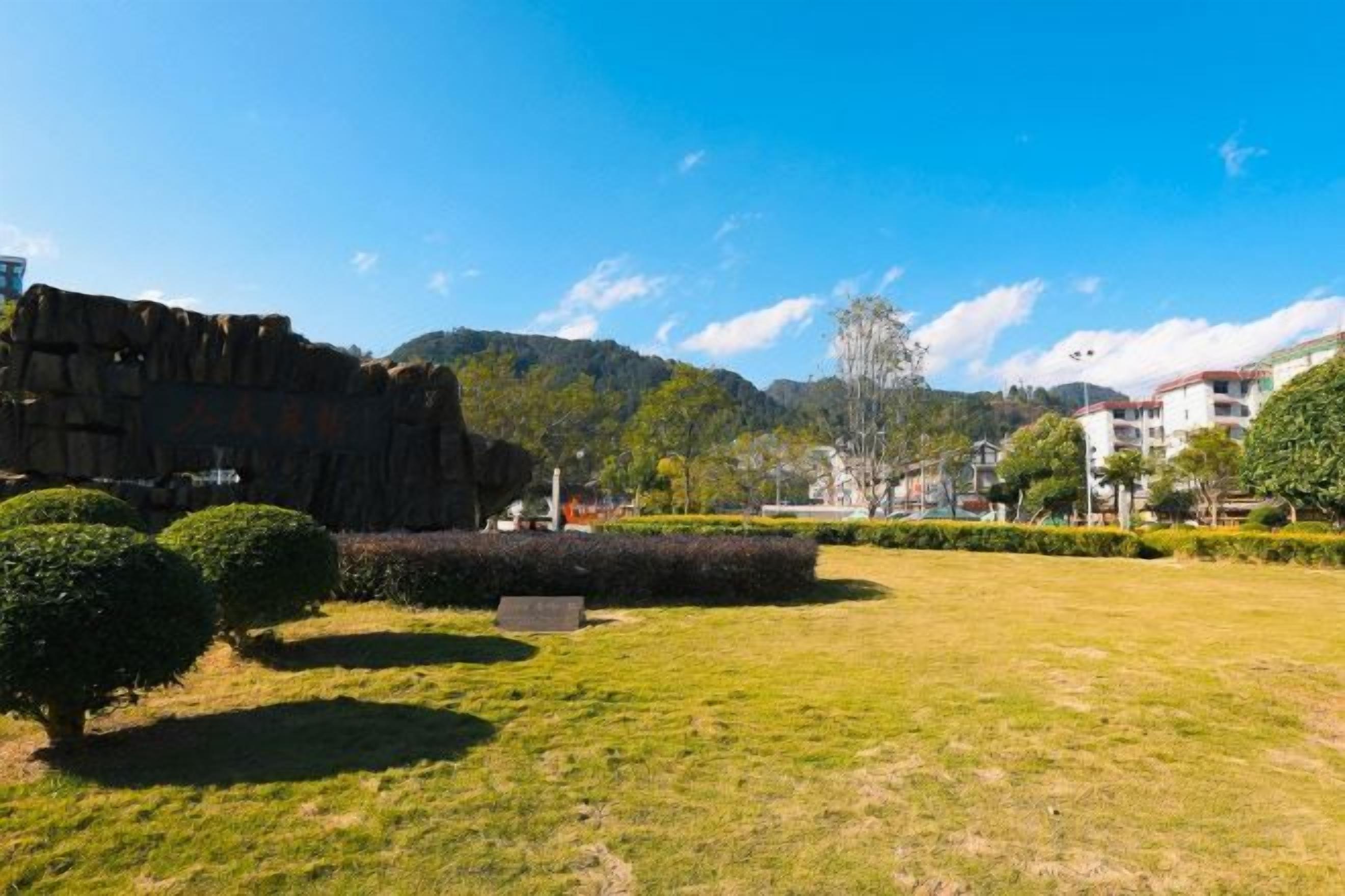 A park with a grassy area, a rock formation, and buildings in the background.