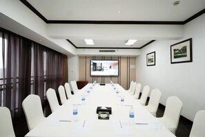 Uma sala de conferências com uma mesa comprida, cadeiras brancas e uma tela de projetor.