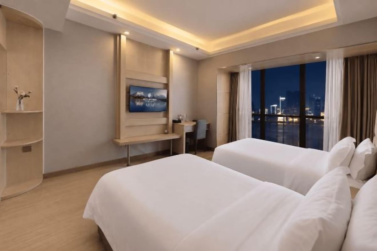A hotel room with two beds, a TV mounted on the wall, a desk, and a view of the cityscape at night.