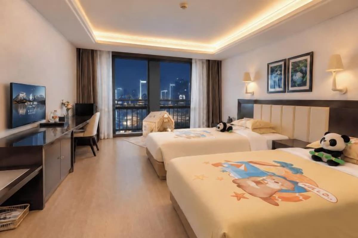 A hotel room with two beds, a desk, a TV, and a balcony with a city view.
