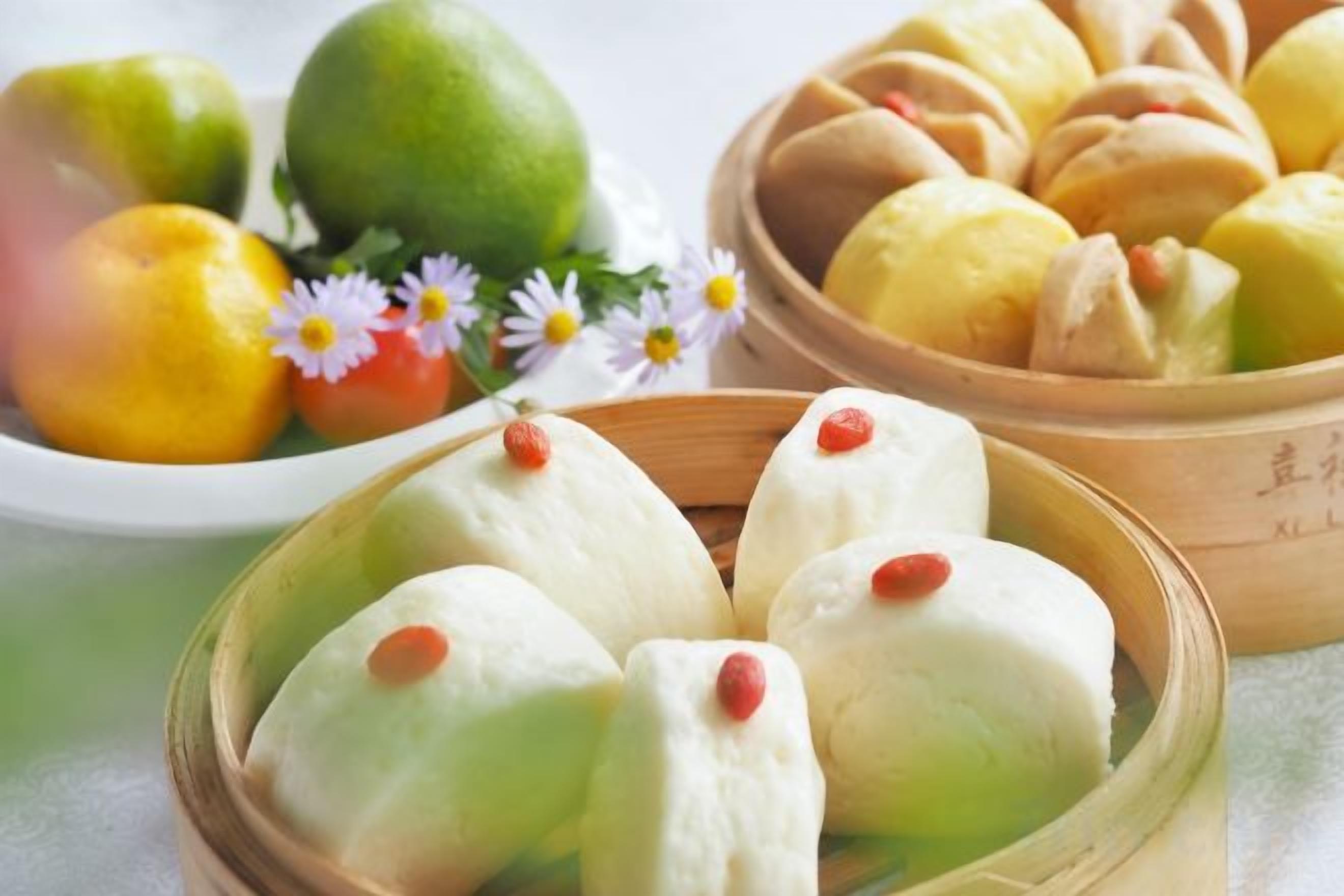 A selection of Asian-style buns in a wooden basket, accompanied by a bowl of fruit and a plate of dumplings.