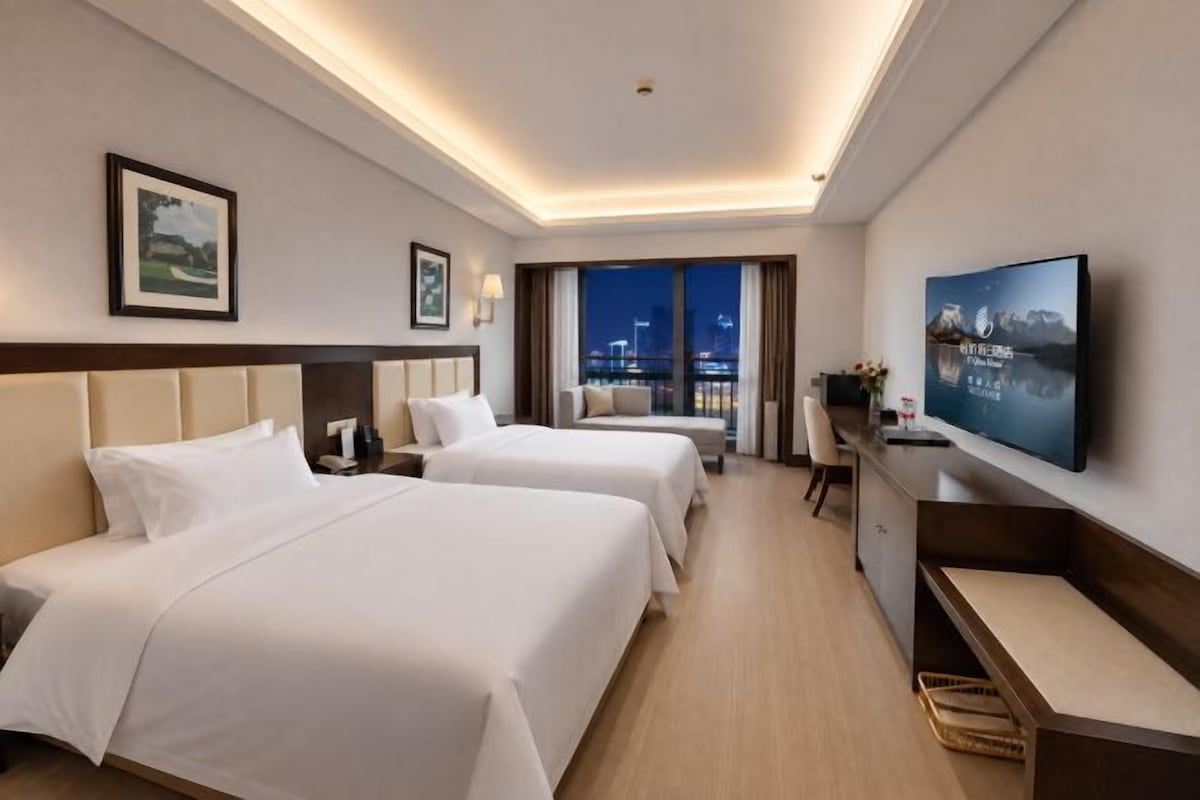 A hotel room with two beds, a desk, a television, and a view of the city at night.