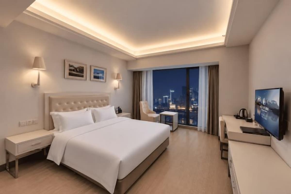 A hotel room with a large bed, bedside tables, a desk, a chair, a TV, and a view of the cityscape.