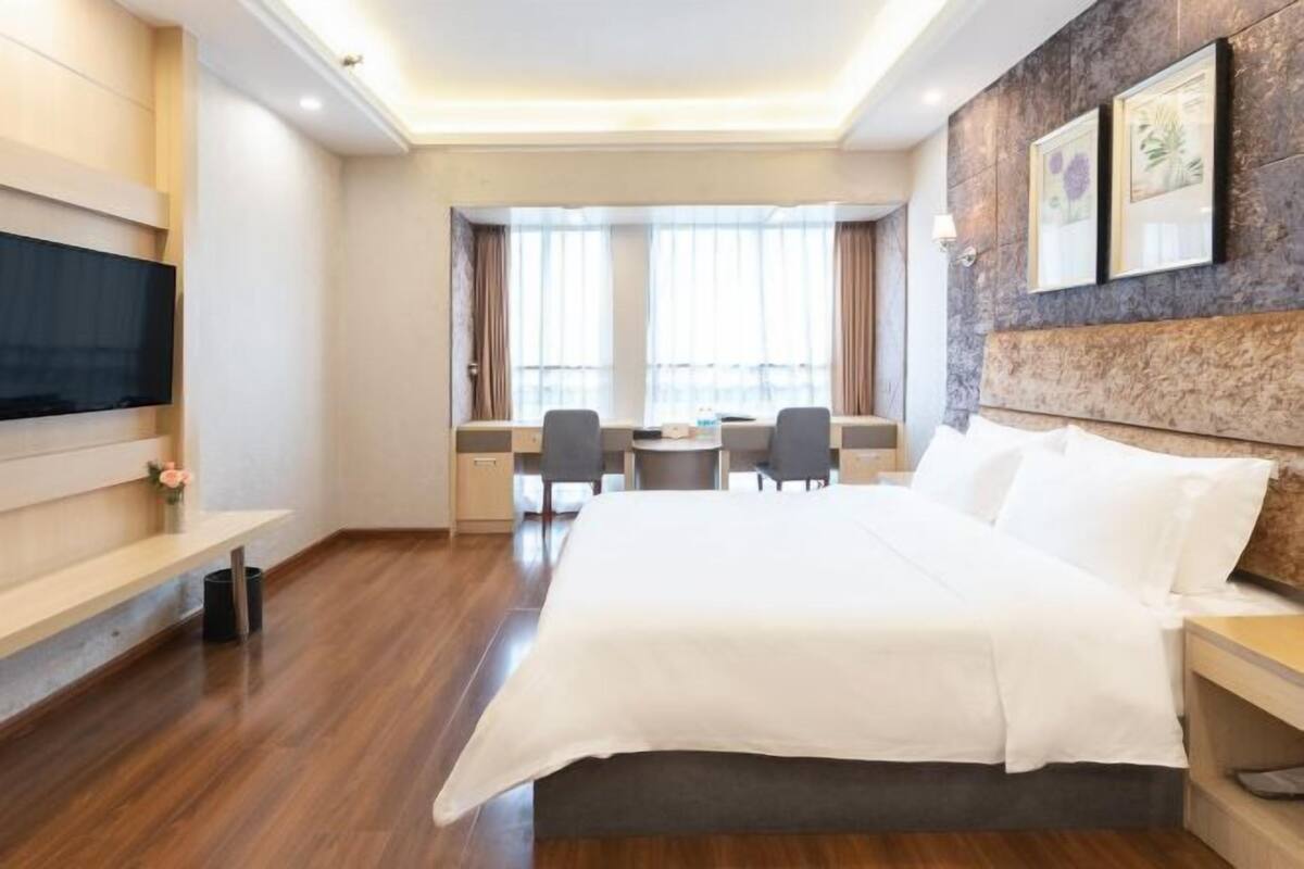 A hotel room with a bed, bedside tables, a desk with chairs, a television, and a large window with curtains.
