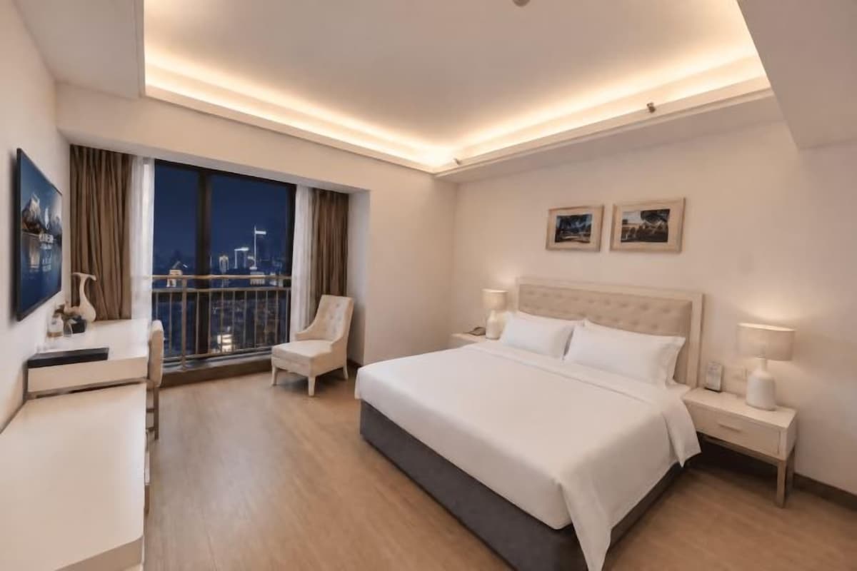 A hotel room with a large bed, bedside tables, a desk, a chair, and a view of the city at night.