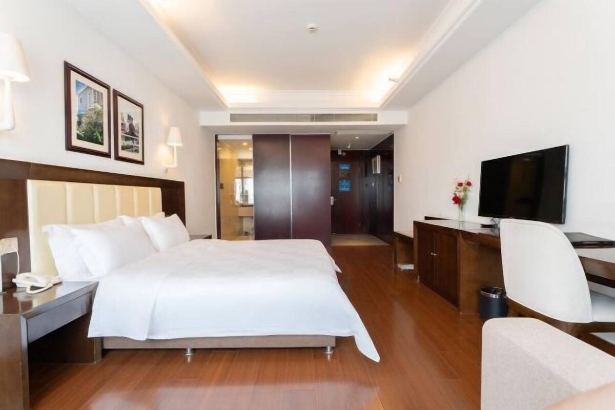 A hotel room with a bed, bedside table, lamp, TV, and a desk with a chair.