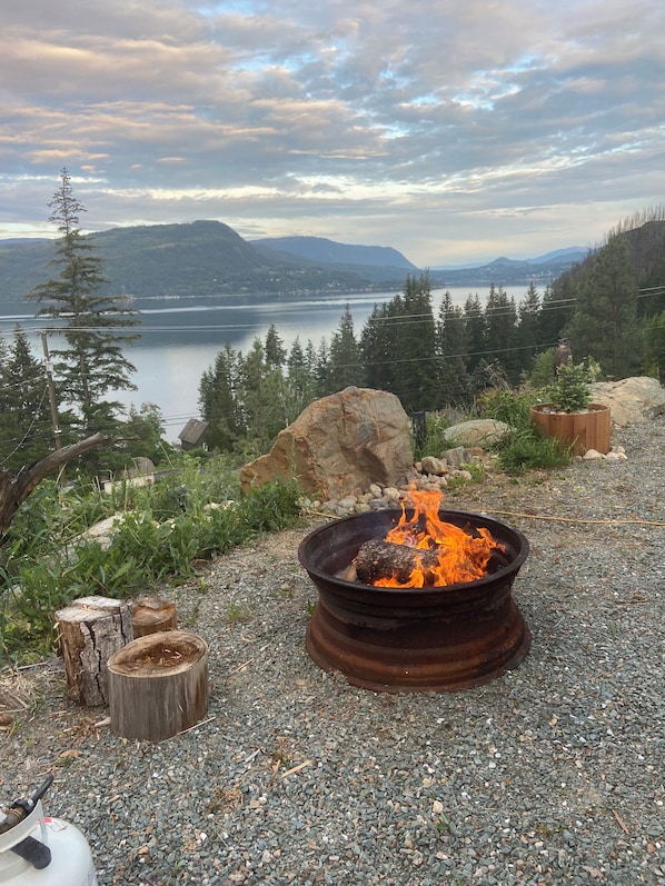 A gravel area with a fire pit, wooden logs, and a view of a lake and mountains.