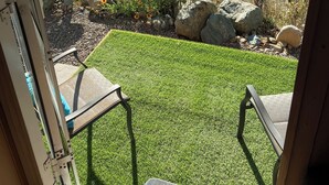 Vista desde una ventana a un patio trasero con césped artificial, un bebedero para pájaros y una zona de patio con sillas.