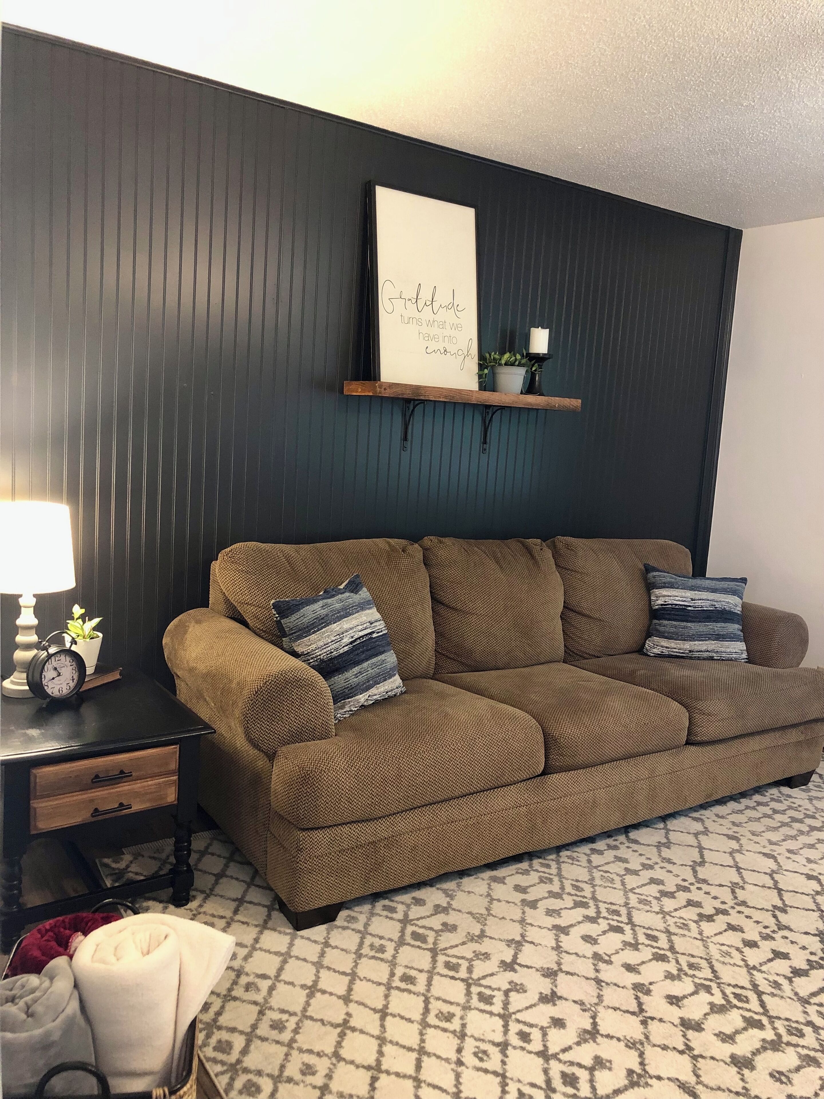 Un salon avec un canapé marron, deux coussins, une petite table sur laquelle se trouvent une horloge et une plante, et une étagère fixée au mur avec un panneau.