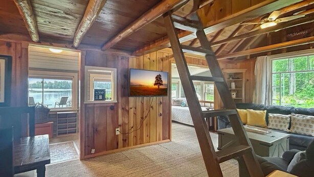 Een gezellig interieur van een chalet met houten plafond, een aan de muur bevestigde televisie, een bank met kussens, en uitzicht op de omgeving door een raam.