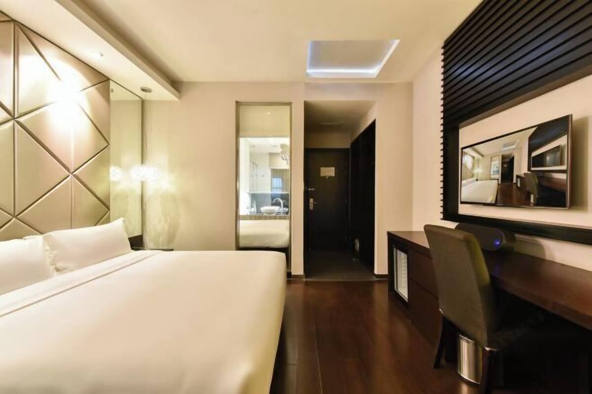 A hotel room with a bed, a desk, a chair, a mirror, and a hallway leading to a bathroom.