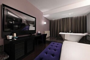 Habitación de hotel moderna con una amplia televisión de pantalla plana, una mesa consola negra con cafetera, un reposapiés púrpura, una cama blanca y un baño independiente con bañera y cortinas.
